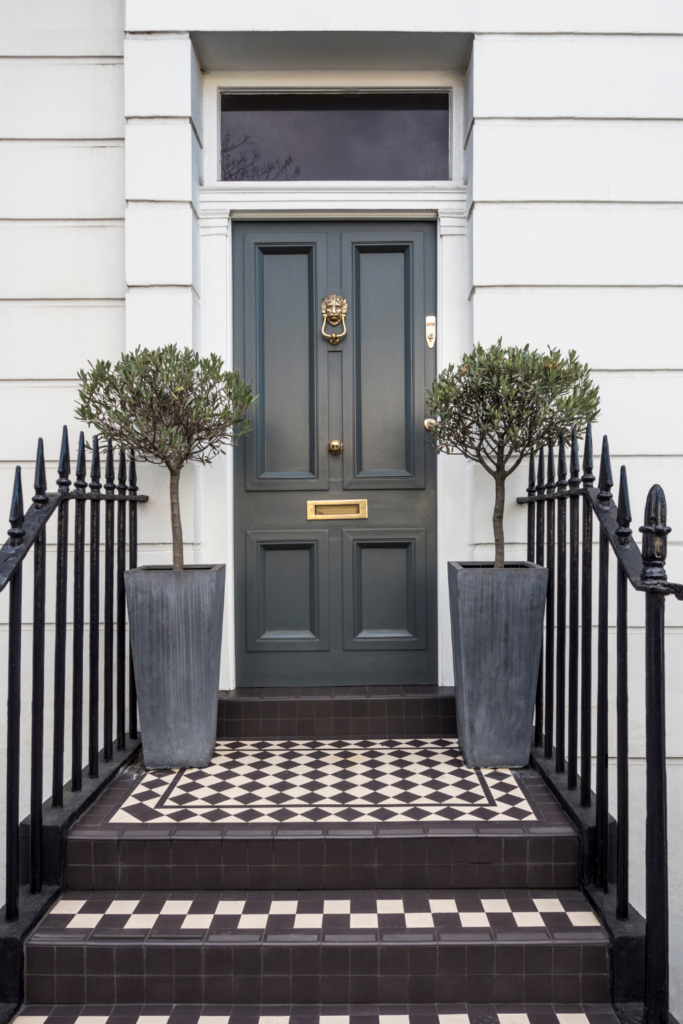 Period style home with Grey front door, two plant pots and black and white tiles. 